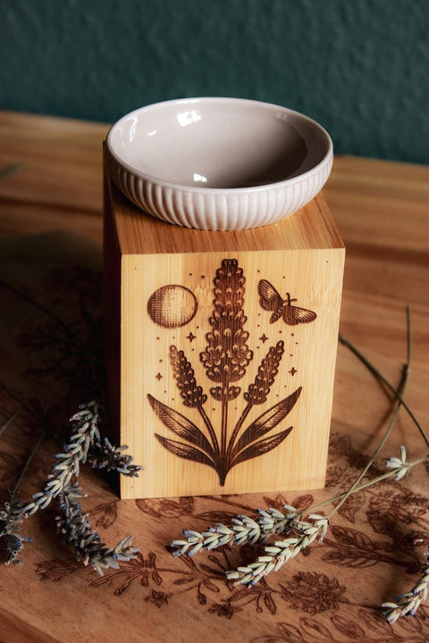 Eine Duftlampe aus Holz und grau-beiger Keramik. In das Holz eingebrannt ist das Motiv eines Lavendels, daneben der Mond, eine Motte und kleine Sterne.
