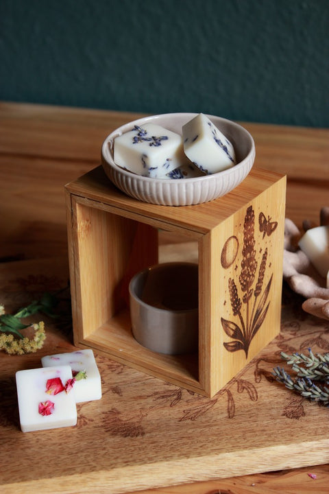 Eine Duftlampe aus Holz und grau-beiger Keramik. In das Holz eingebrannt ist das Motiv eines Lavendels, daneben der Mond, eine Motte und kleine Sterne. In der Keramikschale liegt Duftwachs mit Lavendelblüten.