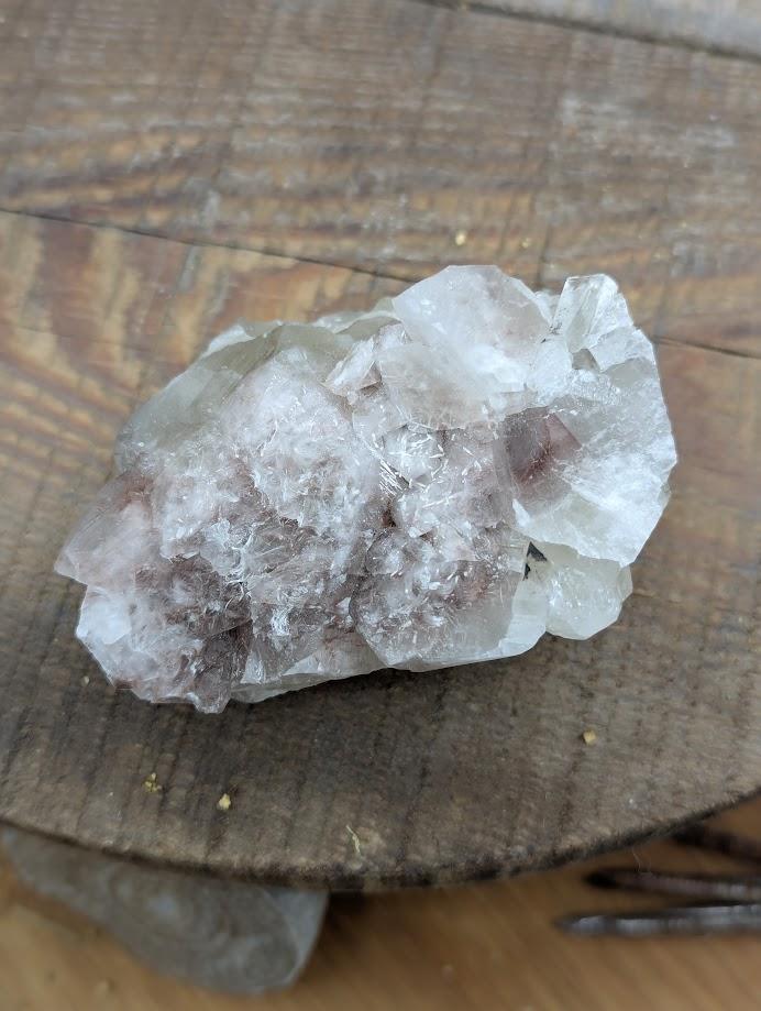 Das Bild zeigt ein Stück Calcit, das auf einer Holzoberfläche liegt. Der Calcit hat eine weiße bis leicht rosa Farbe und besteht aus mehreren kristallinen Strukturen, die in verschiedene Richtungen zeigen. Die Oberfläche des Calcits ist teilweise durchsichtig und weist einige Bruchstellen auf, die die kristalline Struktur des Minerals sichtbar machen.