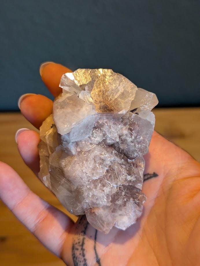 Das Bild zeigt ein Stück Calcit, das in einer Hand gehalten wird. Der Calcit hat eine weiße bis leicht rosa Farbe und besteht aus mehreren kristallinen Strukturen, die in verschiedene Richtungen zeigen. Die Oberfläche des Calcits ist teilweise durchsichtig und weist einige Bruchstellen auf, die die kristalline Struktur des Minerals sichtbar machen.