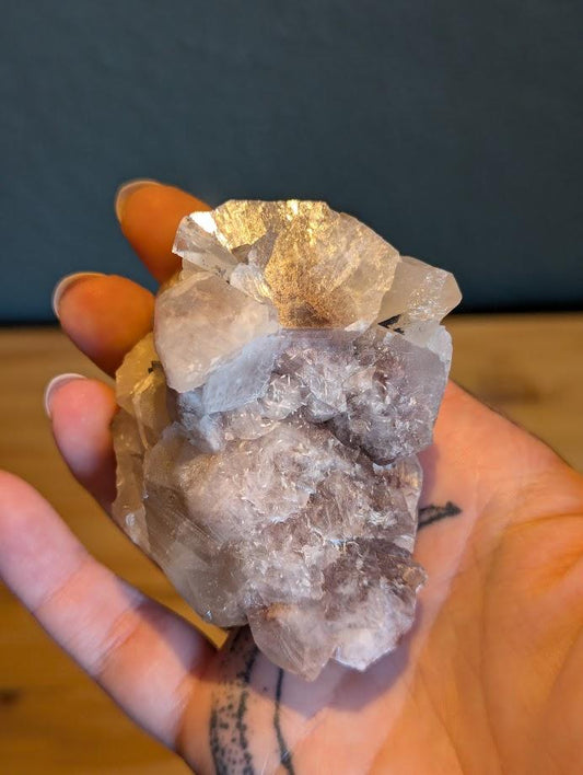 Das Bild zeigt ein Stück Calcit, das in einer Hand gehalten wird. Der Calcit hat eine weiße bis leicht rosa Farbe und besteht aus mehreren kristallinen Strukturen, die in verschiedene Richtungen zeigen. Die Oberfläche des Calcits ist teilweise durchsichtig und weist einige Bruchstellen auf, die die kristalline Struktur des Minerals sichtbar machen.