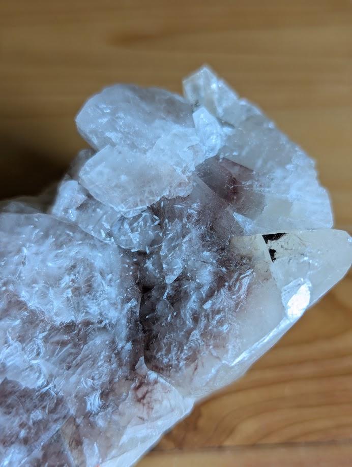 Das Bild zeigt ein Stück Calcit, das auf einer Holzoberfläche liegt. Der Calcit hat eine weiße bis leicht rosa Farbe und besteht aus mehreren kristallinen Strukturen, die in verschiedene Richtungen zeigen. Die Oberfläche des Calcits ist teilweise durchsichtig und weist einige Bruchstellen auf, die die kristalline Struktur des Minerals sichtbar machen.