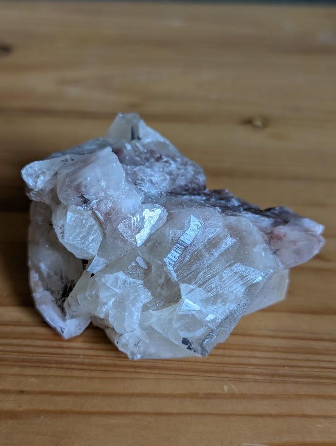 Das Bild zeigt ein Stück Calcit, das auf einer Holzoberfläche liegt. Der Calcit hat eine weiße bis leicht rosa Farbe und besteht aus mehreren kristallinen Strukturen, die in verschiedene Richtungen zeigen. Die Oberfläche des Calcits ist teilweise durchsichtig und weist einige Bruchstellen auf, die die kristalline Struktur des Minerals sichtbar machen.