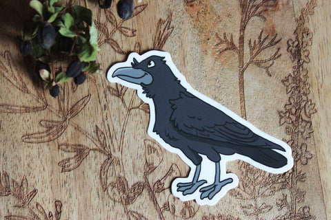 Das Bild zeigt einen Aufkleber auf einer Holzoberfläche mit kunstvollen Pflanzenmustern. Der Aufkleber selbst stellt einen schwarzen Vogel dar, einen Raben, der detailliert gestaltet ist, mit einem auffälligen Schnabel, Federn und Füßen. Der Vogel hat eine weiße Umrandung, die ihn hervorhebt. In der oberen linken Ecke des Bildes sind außerdem getrocknete dunkle Beeren zu sehen.
