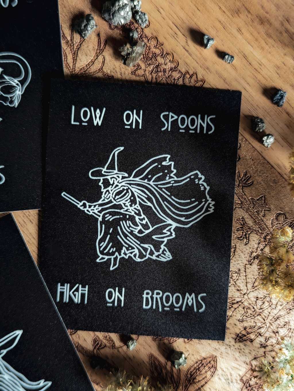 Auf einem Holzbrett liegt ein mattschwarzer Sticker. In weißer Farbe ist eine Hexe auf einem Besen zu sehen. Dazu der Spruch: Low in Spoons High in Brooms