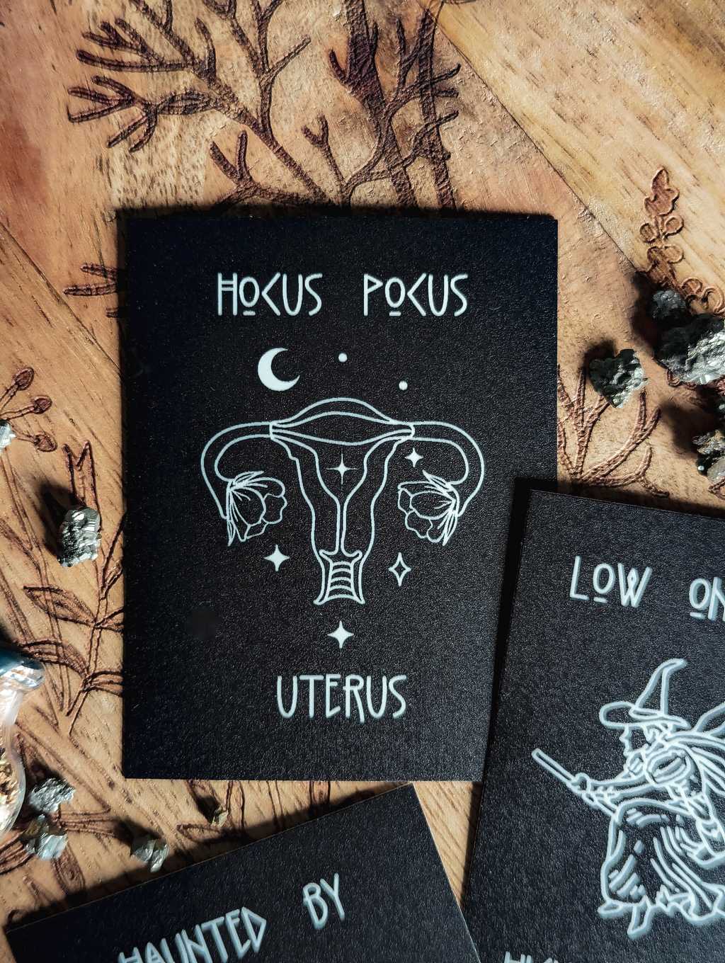 Auf einem Holzbrett liegt ein mattschwarzer Sticker. In weißer Farbe ist ein Uterus mit Blüten, Sternen und Mond zu sehen. Dazu der Spruch: Hocus Pocus Uterus