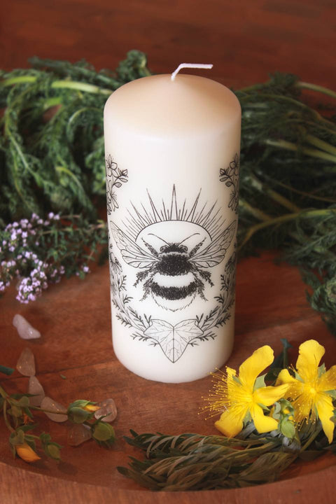 Eine cremeweiße Stumpenkerze mit schwarzem Motiv. Es zeigt eine Hummel. Eingerahmt wird sie von Johanniskraut, Kamille und einem Efeublatt.