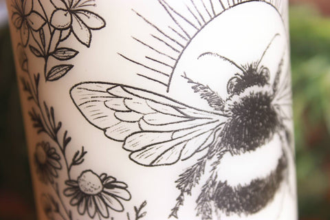 Nahaufnahme: Eine cremeweiße Stumpenkerze mit schwarzem Motiv. Es zeigt eine Hummel. Eingerahmt wird sie von Johanniskraut, Kamille und einem Efeublatt.