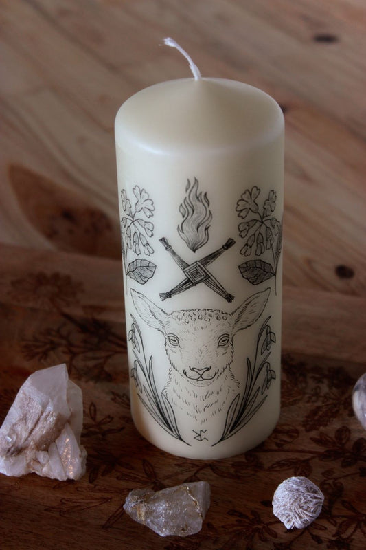 Eine creme-farbene Stumpen-Kerze mit schwarzem Motiv. Es zeigt im Zentrum ein Schaf. Eingerahmt wird es vom frühblühenden Schneeglöckchen und der Schlüsselblume, mit der die Holle in manchen Geschichten das Tor zum Frühling aufschließt. Typische Imbolc-Motive sind die Kerzenflamme und das Kreuz der Brigid.