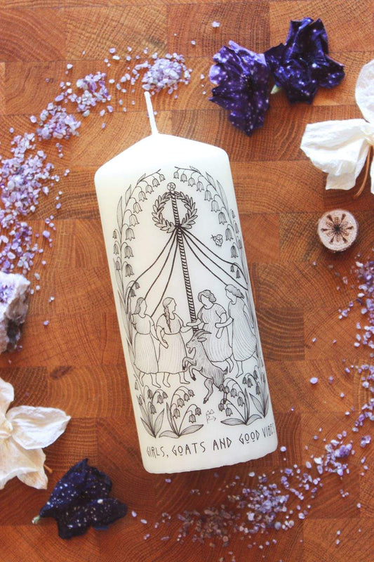 Creme-farbene Stumpenkerze aus veganem Wachs mit schwarzer Linienillustration: Mehrere Frauen und ein Ziegenbock tanzen um einen Maibaum mit flatternden Bändern, umgeben von detaillierten Maiglöckchen‑Ornamenten. Das folkloristische Frühlingsmotiv verbindet Tanz in den Mai, feministische Symbolik und modernen Line‑Art‑Stil. Unten auf der Kerze steht: Girls, Goats and Good Vibes