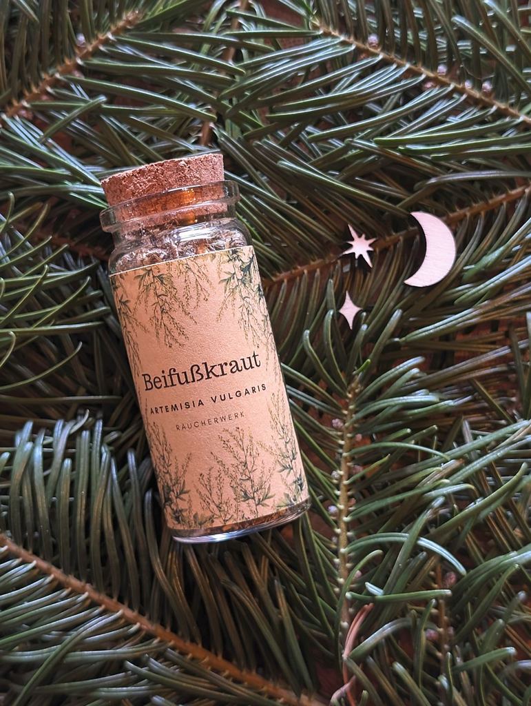 Eine Korkenflasche liegt gebettet auf Tannenzweigen. In der Flasche ist Beifuß zu sehen. Darauf ist ein Etikett mit der Aufschrift: Beifußkraut Artemisia vulgaris Räucherwerk