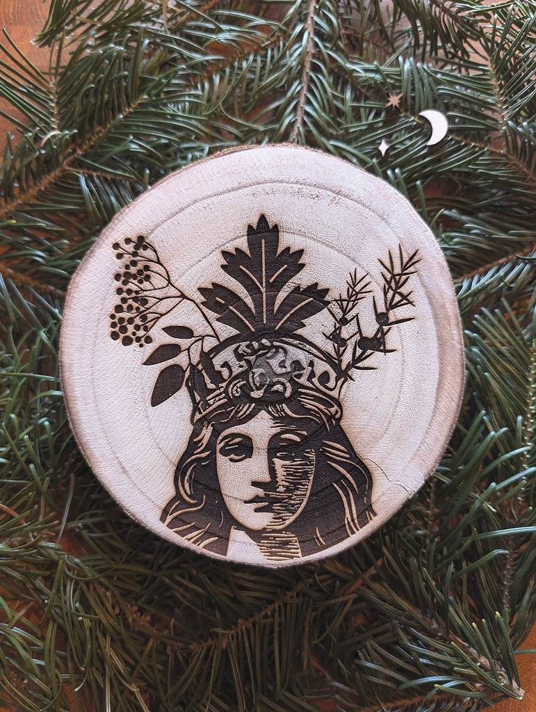Eine große dicke Holzscheibe mit eingraviertem Bild. Es zeigt eine Frau, deren linke Gesichtshälfte im Schatten verdeckt ist. Sie trägt eine Krone aus Beifuß, Wacholder und Holunder.