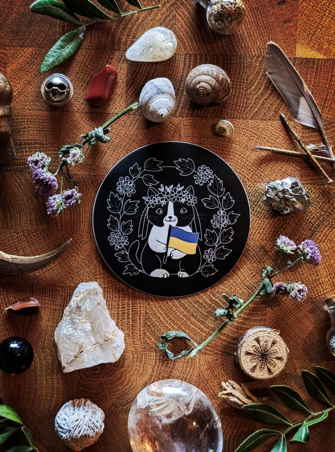 Ein runder schwarzer Sticker mit Motiv. Das Motiv zeigt eine schwarz-weiße niedliche Katze mit Blumenkrone auf dem Kopf. Sie hält die blau-gelbe Flagge der Ukraine in der Pfote. Umrahmt wird sie vom Schneeball-Strauch.