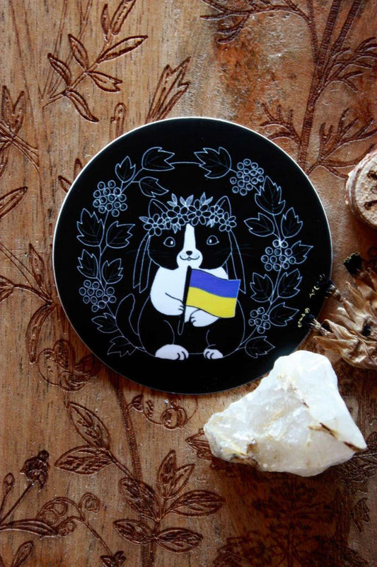 Ein runder schwarzer Sticker mit Motiv. Das Motiv zeigt eine schwarz-weiße niedliche Katze mit Blumenkrone auf dem Kopf. Sie hält die blau-gelbe Flagge der Ukraine in der Pfote. Umrahmt wird sie vom Schneeball-Strauch.