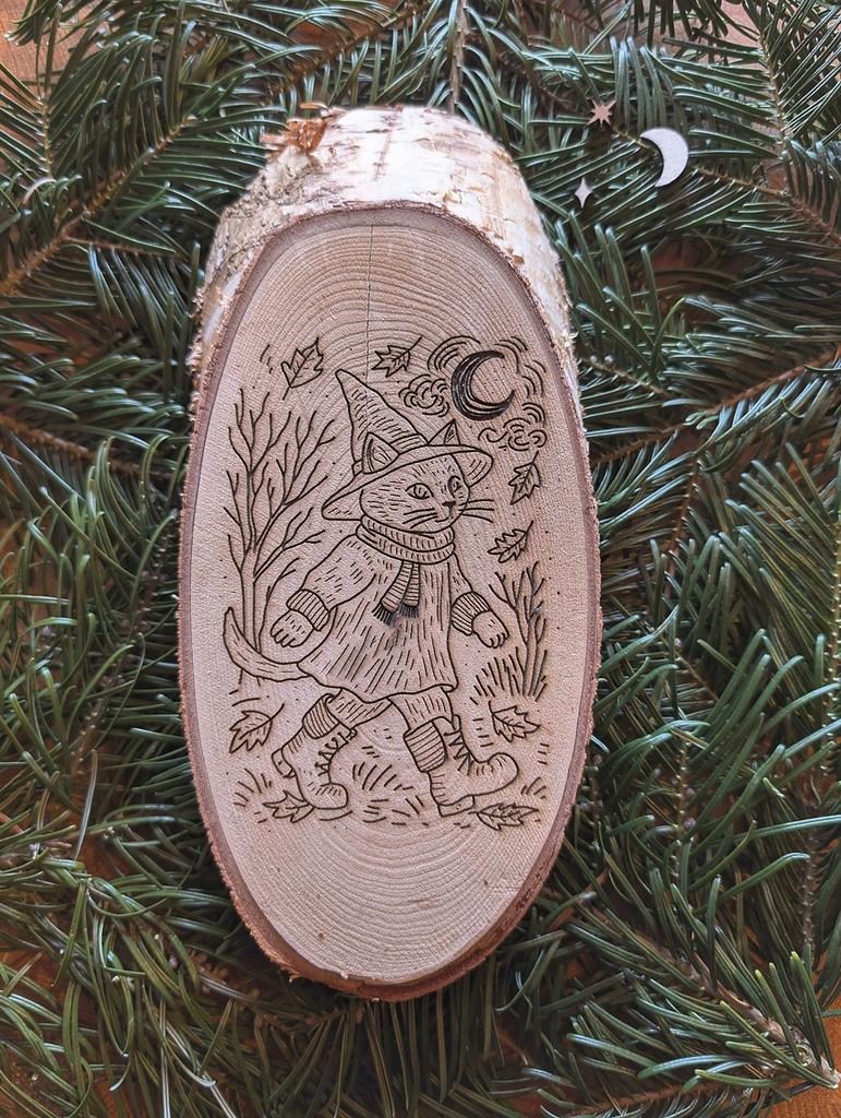 Auf Tannenzweigen liegt eine ovale Birkenholzscheibe. Die rustikale Baumscheibe aus Birkenholz zeigt eine Katze mit Stiefelchen, Pullover, Schal und Hexenhut, die gerade durch den Herbst spaziert.