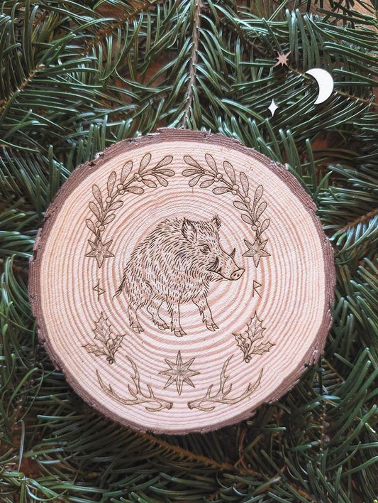 Gebettet auf Tannenzweigen liegt in der Mitte eine runde Scheibe aus Holz. Diese Dekoration zu Jul (Wintersonnenwende) zeigt ein Wildschwein. Eingerahmt wird es von typischen Jul-Motiven wie Mistelzweigen, Stechpalmen, Strohsternen, Geweihen und passenden Runen.