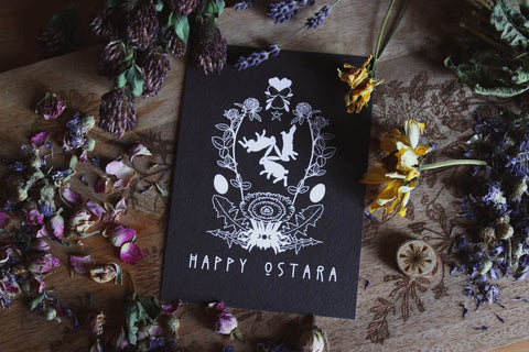 Das Bild zeigt eine schwarze Karte mit weißen Illustrationen und dem Text „HAPPY OSTARA“ unten. Um die Karte herum liegen verschiedene getrocknete Blumen und Kräuter, darunter Klee, Lavendel und Rosen. Auf der Karte sind drei Hasen, Eier und florale Elemente in einem symmetrischen Design abgebildet. Die Karte liegt auf einer Holzoberfläche mit weiteren floralen Gravuren.