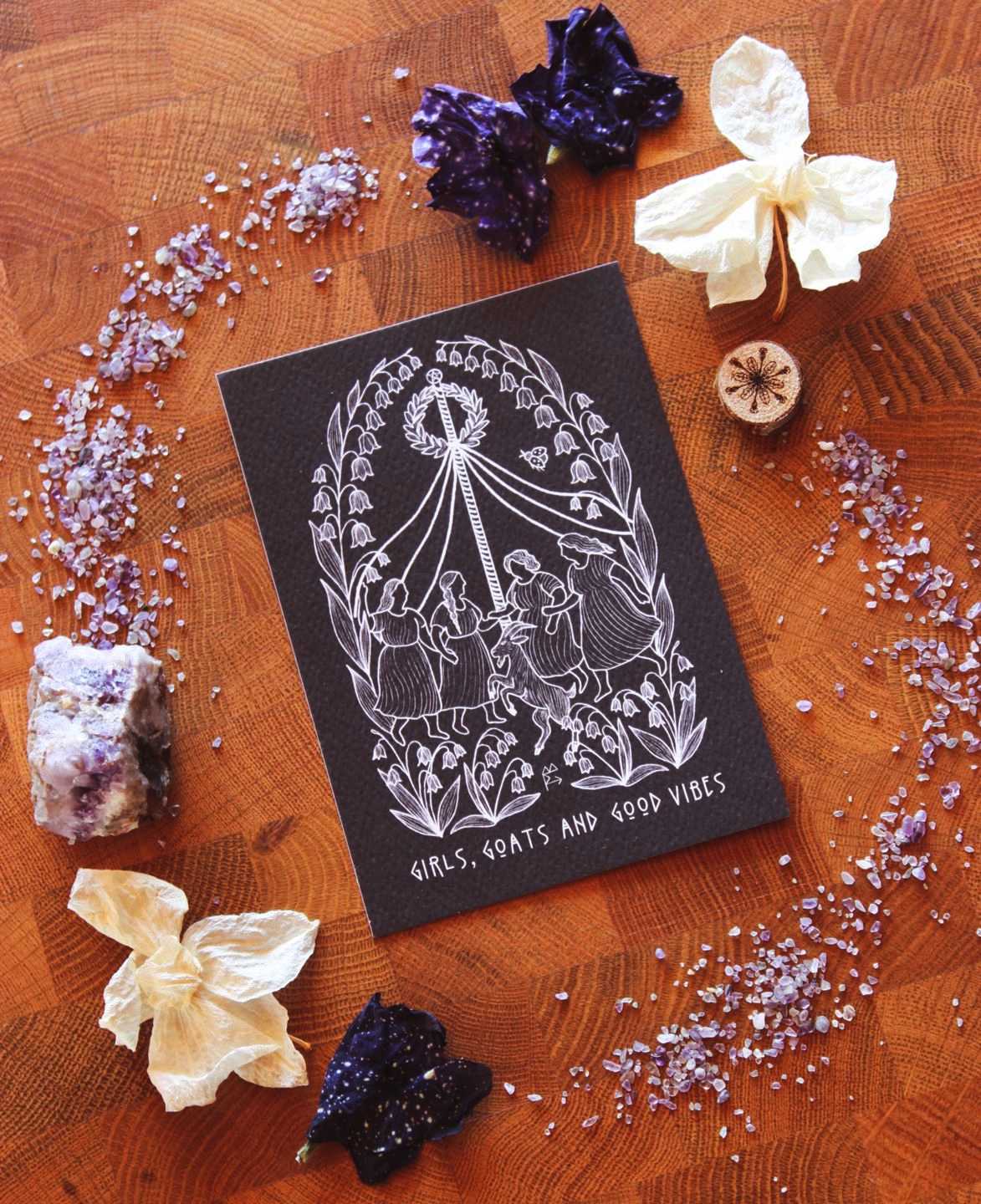 Schwarzer Print mit weißer Linienillustration: Mehrere Frauen und ein Ziegenbock tanzen um einen Maibaum mit flatternden Bändern, eingerahmt von detaillierten Maiglöckchen‑Ornamenten. Das Motiv erinnert an folkloristische Frühlingsrituale und verbindet tanzende Figuren, Natur und symbolische Maiglöckchen in einem klaren, modernen Line‑Art‑Stil.
Unten auf dem Print steht: Girls, Goats and Good Vibes