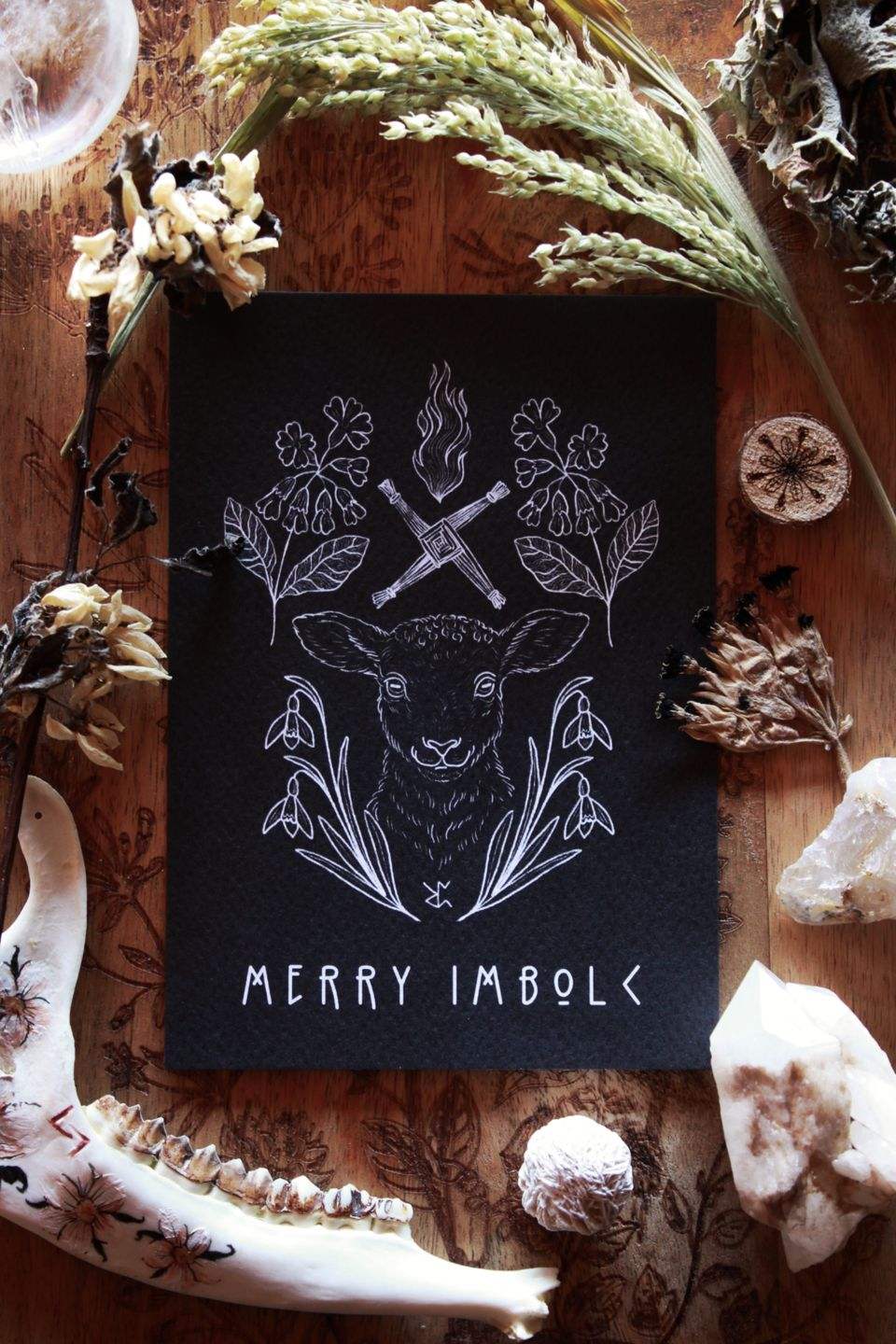 Diese schwarze Grußkarte mit weißem Druck zu Imbolc zeigt im Zentrum ein Schaf. Eingerahmt wird es vom frühblühenden Schneeglöckchen und der Schlüsselblume, mit der die Holle in manchen Geschichten das Tor zum Frühling aufschließt. Typische Imbolc-Motive sind die Kerzenflamme und das Kreuz der Brigid. Unten auf der Karte steht: Merry Imbolc