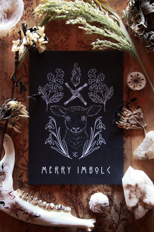 Diese schwarze Grußkarte mit weißem Druck zu Imbolc zeigt im Zentrum ein Schaf. Eingerahmt wird es vom frühblühenden Schneeglöckchen und der Schlüsselblume, mit der die Holle in manchen Geschichten das Tor zum Frühling aufschließt. Typische Imbolc-Motive sind die Kerzenflamme und das Kreuz der Brigid. Unten auf der Karte steht: Merry Imbolc