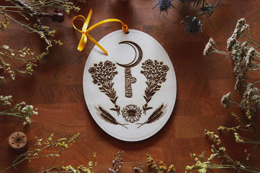 Dieses Holz-Ornament zu Lammas (Lughnasadh / Schnitterfest) zeigt klassische Motive für das erste der drei Erntefeste im Jahreskreis. Die Sichel ist von zwei Sträußen Flachs (Lein) umgeben. Eine Pflanze die fest in Verbindung zur Frau Holle steht. Darunter ist eine Sonnenblumenblüte und daneben vier Roggenähren. Das Band ist in einem kräftigen Gelb gehalten.