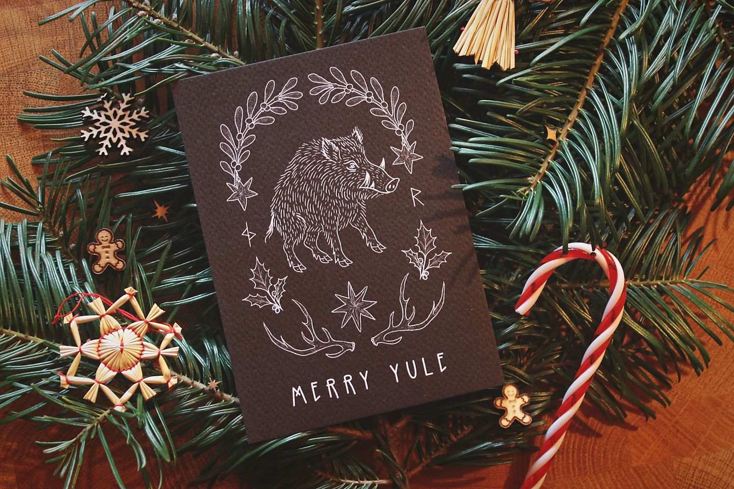 Gebettet auf Tannenzweigen und weihnachtliche Dekoration liegt in der Mitte eine schwarze Karte mit weißem Aufdruck. Diese Grußkarte zu Jul (Wintersonnenwende) zeigt ein Wildschwein. Eingerahmt wird es von typischen Jul-Motiven wie Mistelzweigen, Stechpalmen, Strohsternen, Geweihen und passenden Runen.