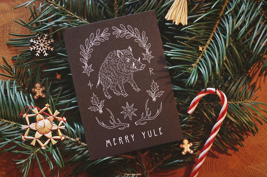 Gebettet auf Tannenzweigen und weihnachtliche Dekoration liegt in der Mitte eine schwarze Karte mit weißem Aufdruck. Diese Grußkarte zu Jul (Wintersonnenwende) zeigt ein Wildschwein. Eingerahmt wird es von typischen Jul-Motiven wie Mistelzweigen, Stechpalmen, Strohsternen, Geweihen und passenden Runen.