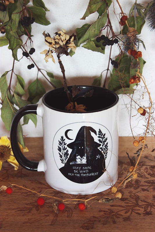 Die Tasse aus Keramik trägt das Motiv "Stay Safe Be Kind Hex The Patriarchy". Die vermummte Hexen-Gestalt mit Hexen-Hut, hält eine Katze in den Armen. Auf ihrer Schulter ist ein kleines Pentagramm zu sehen. Um sie herum sind Salbei und Beifuß. Das Motiv ist in schwarz gehalten, genau so wie der Henkel und das Innere der Tasse. Ansonsten ist die Tasse weiß. Umrahmt wird die Tasse von vielen Pflanzen.