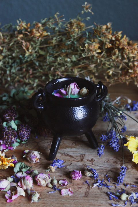 Das Bild zeigt einen kleinen schwarzen Kessel, der auf einer Holzoberfläche steht. Umgeben ist der Kessel von verschiedenen getrockneten Blumen und Kräutern, darunter rosafarbene Rosenknospen, gelbe und lila Blüten. Innerhalb des Kessels befinden sich einige getrocknete Rosenknospen. Im Hintergrund sind weitere getrocknete Pflanzen und Kräuter verstreut, was eine rustikale und natürliche Atmosphäre schafft.