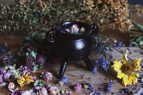 Das Bild zeigt einen kleinen schwarzen Kessel, der auf einer Holzoberfläche steht. Umgeben ist der Kessel von verschiedenen getrockneten Blumen und Kräutern, darunter rosafarbene Rosenknospen, gelbe und lila Blüten. Innerhalb des Kessels befinden sich einige getrocknete Rosenknospen. Im Hintergrund sind weitere getrocknete Pflanzen und Kräuter verstreut, was eine rustikale und natürliche Atmosphäre schafft.