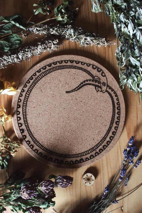 Das Bild zeigt eine Korkunterlage mit dem schwarzen Symbol einer sich selbst verschlingenden Schlange, bekannt als Ouroboros. Sie liegt auf einer Holzfläche und ist von verschiedenen getrockneten Kräutern und Blumen umgeben, darunter Lavendel, Salbei und weitere botanische Elemente.