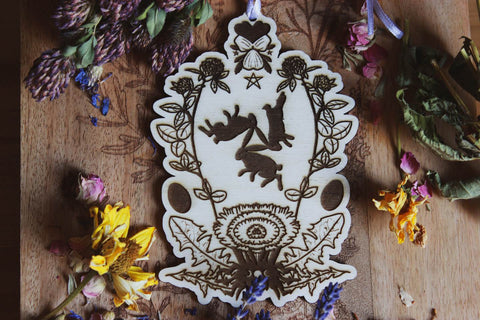 Das Bild zeigt ein kunstvolles Holzornament mit detailliertem Motiv und einer fliederfarbenen Schleife, die oben gebunden ist. Im Zentrum des Ornaments sind drei Hasen dargestellt, die im Kreis laufen, umgeben von floralen Mustern, wie Rotklee, Löwenzahn und Veilchen. Das Ornament liegt auf einer Holzoberfläche, umgeben von verschiedenen getrockneten Blumen, wie Kleeblüten, gelben Blumen und Lavendel.