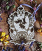Das Bild zeigt ein kunstvolles Holzornament mit detailliertem Motiv und einer fliederfarbenen Schleife, die oben gebunden ist. Im Zentrum des Ornaments sind drei Hasen dargestellt, die im Kreis laufen, umgeben von floralen Mustern, wie Rotklee, Löwenzahn und Veilchen. Das Ornament liegt auf einer Holzoberfläche, umgeben von verschiedenen getrockneten Blumen, wie Kleeblüten, gelben Blumen und Lavendel.
