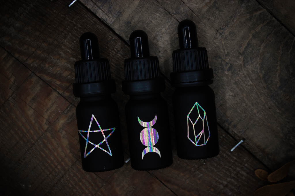 Drei mattschwarze Pipettenflaschen mit drei Symbolen in changierenden Tönen: Pentagramm, Dreifachmond und Kristall.