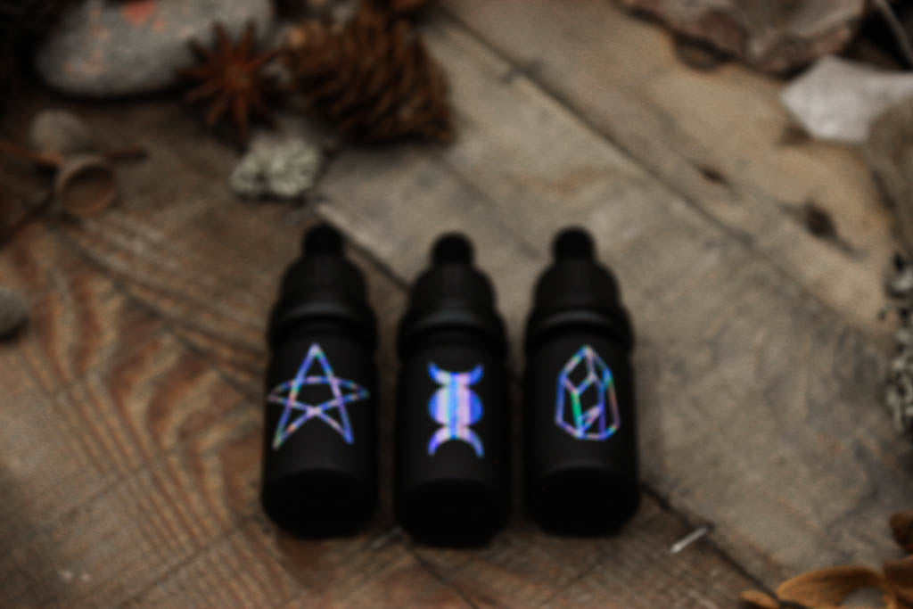 Drei mattschwarze Pipettenflaschen mit drei Symbolen in changierenden Tönen: Pentagramm, Dreifachmond und Kristall. Das Bild ist unscharf um den multichrome Effekt besser zu verdeutlichen.