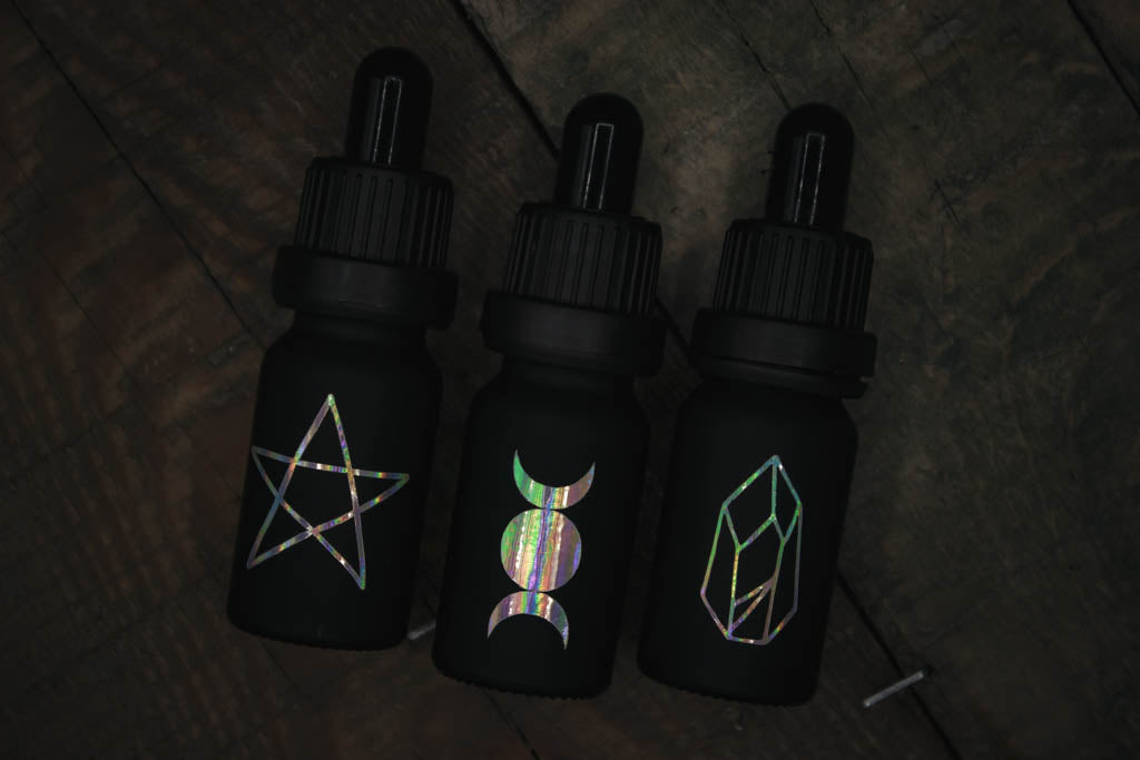 Drei mattschwarze Pipettenflaschen mit drei Symbolen in changierenden Tönen: Pentagramm, Dreifachmond und Kristall.