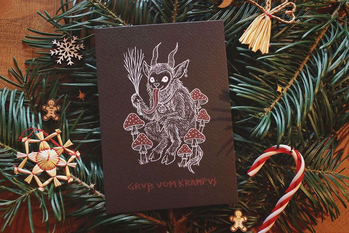 Gebettet auf Tannenzweigen und weihnachtlicher Dekoration liegt in der Mitte eine schwarze Karte mit weiß-rotem Aufdruck. Das Motiv zeigt einen Krampus, der zwischen Fliegenpilzen sitzt. Er hat einen Pentagramm-Ohrring am Ohr, eine große Glocke um den Hals und hält eine Rute aus Zweigen in der Klaue. In roter Schrift steht darunter: Gruß vom Krampus