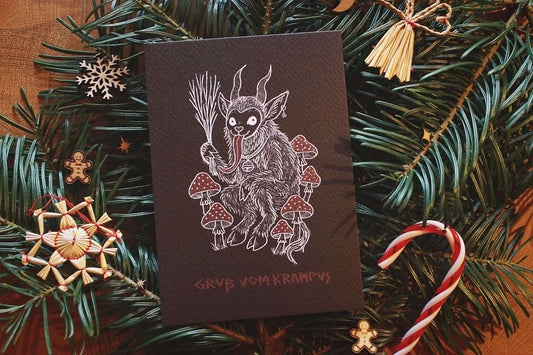 Gebettet auf Tannenzweigen und weihnachtlicher Dekoration liegt in der Mitte eine schwarze Karte mit weiß-rotem Aufdruck. Das Motiv zeigt einen Krampus, der zwischen Fliegenpilzen sitzt. Er hat einen Pentagramm-Ohrring am Ohr, eine große Glocke um den Hals und hält eine Rute aus Zweigen in der Klaue. In roter Schrift steht darunter: Gruß vom Krampus