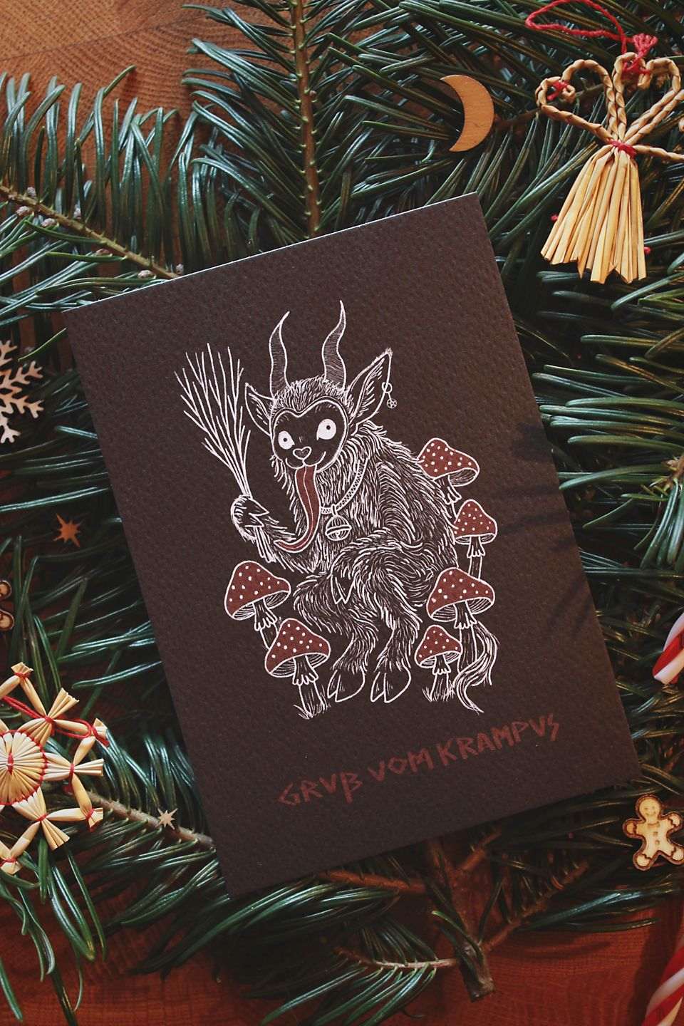 Hochkant: Gebettet auf Tannenzweigen und weihnachtlicher Dekoration liegt in der Mitte eine schwarze Karte mit weiß-rotem Aufdruck. Das Motiv zeigt einen Krampus, der zwischen Fliegenpilzen sitzt. Er hat einen Pentagramm-Ohrring am Ohr, eine große Glocke um den Hals und hält eine Rute aus Zweigen in der Klaue. In roter Schrift steht darunter: Gruß vom Krampus