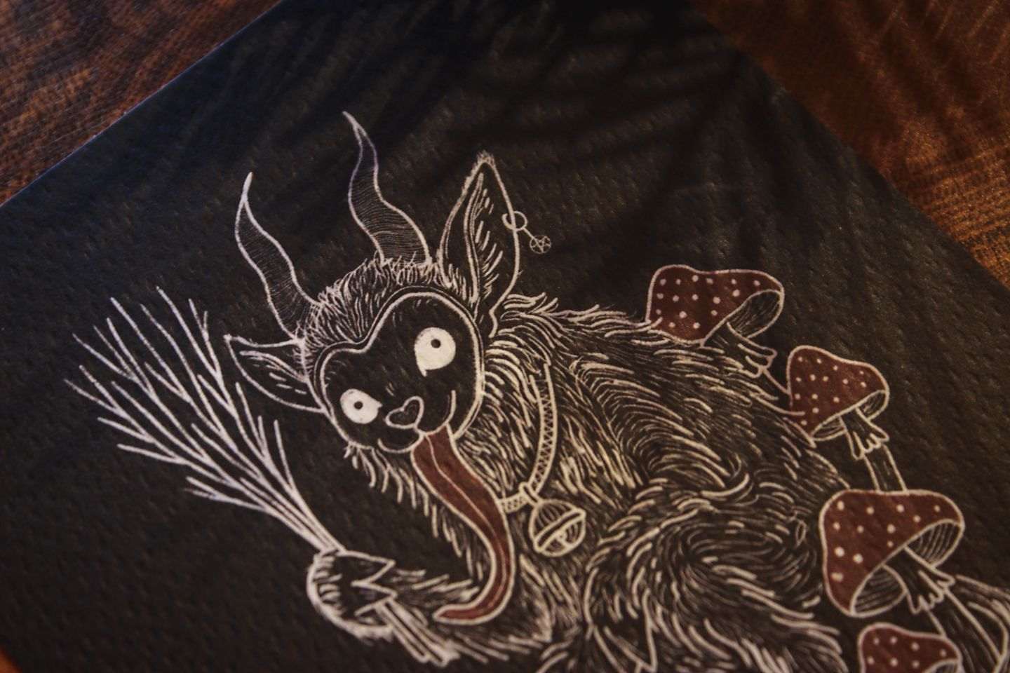 Nahaufnahme: Gebettet auf Tannenzweigen und weihnachtlicher Dekoration liegt in der Mitte eine schwarze Karte mit weiß-rotem Aufdruck. Das Motiv zeigt einen Krampus, der zwischen Fliegenpilzen sitzt. Er hat einen Pentagramm-Ohrring am Ohr, eine große Glocke um den Hals und hält eine Rute aus Zweigen in der Klaue. In roter Schrift steht darunter: Gruß vom Krampus