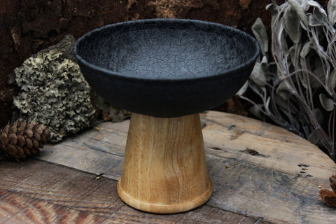 Auf einer runden Holzplatte steht ein schlicht-eleganter und moderner Räucher-Kelch. Sein Körper ist aus schwarzem pulverbeschichteten Eisen. Und sein Fuß ist aus Mangoholz.
