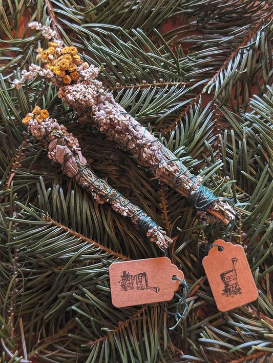 Ein kleiner und ein großer Räucherbündel liegen auf Tannenzweigen nebeneinander. An ihnen ist ein Kartonschild mit dem Motiv eines Mörsers.