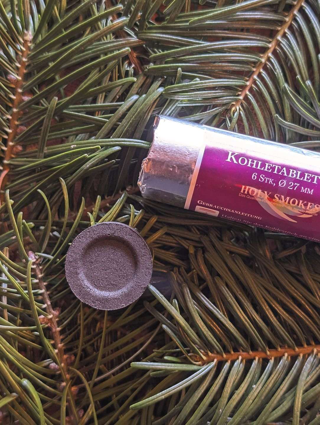 Eine Rolle Kohletabletten zum Räuchern liegen auf Tannenzweigen. Daneben liegt ein einzelnes Stück Räucherkohle.