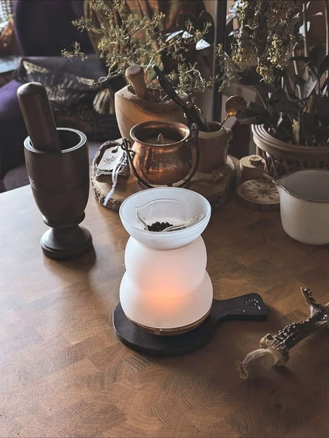 Ein rustikales, gemütliches Arrangement auf einem Holztisch mit einem beleuchteten Räucher-Stövchen aus Milchglas in dem dunkle Beeren liegen, einem dunklen Mörser mit Stößel, einem Kupferkessel und getrockneten Kräutern. Dekorative Elemente wie ein Holztablett, ein kleines Keramikgefäß und ein geschnitztes Geweih ergänzen die Szene. Im Hintergrund sind Pflanzen und ein mit Stoff bezogener Stuhl zu sehen – eine warme, naturverbundene Atmosphäre.