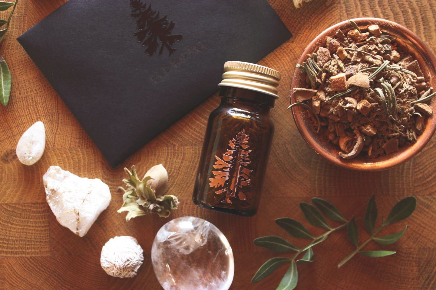 Flach gelegte Szene mit einem kleinen Braunglasfläschchen mit Baumemblem, einer Schale voller getrocknetem Räucherwerk und verschiedenen Naturmaterialien wie Steinen, Nüssen und Blättern auf einem Holzhintergrund.