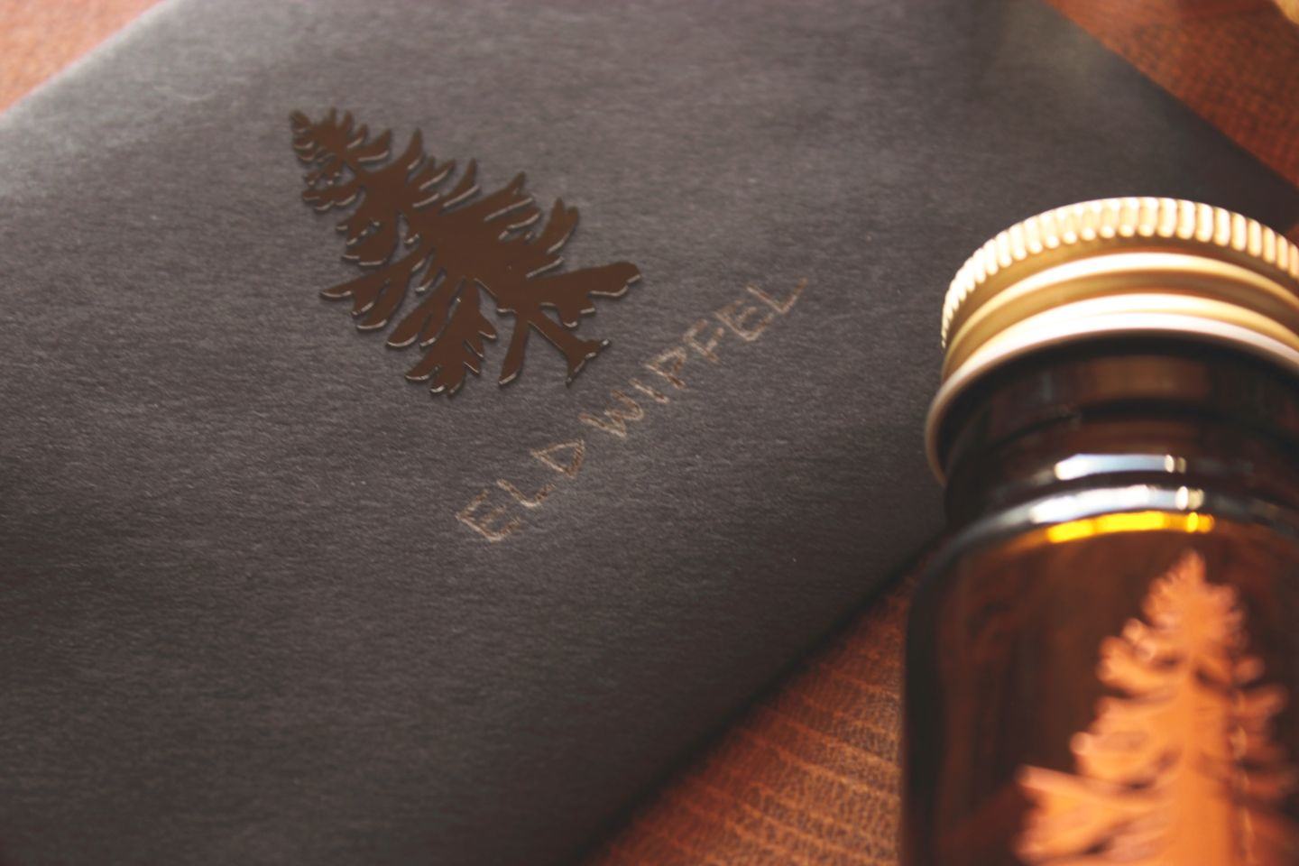 Nahaufnahme eines dunklen Umschlags und eines Braunglasgefäßes mit goldenem Deckel, beide mit glänzendem Baumlogo; im Gefäß befindet sich Räucherwerk.