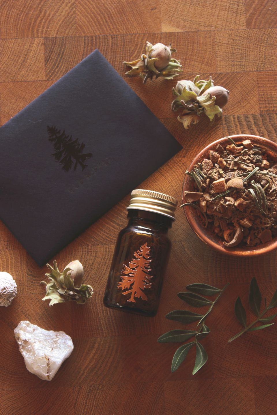 Flach gelegte Szene mit einem kleinen Braunglasfläschchen mit Baumemblem, einer Schale voller getrocknetem Räucherwerk und verschiedenen Naturmaterialien wie Steinen, Nüssen und Blättern auf einem Holzhintergrund.