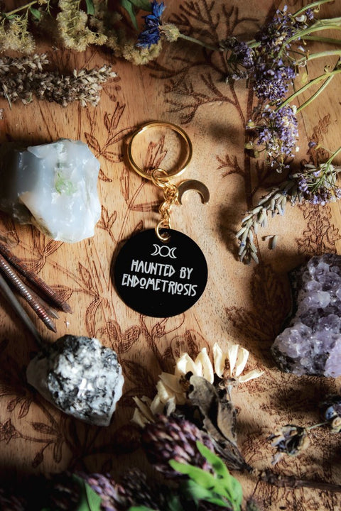 Ein Schlüsselanhänger in Schwarz und Gold mit goldenem Mond-Charm. Auf der schwarzen Scheibe steht in weiß: Haunted by Endometriosis