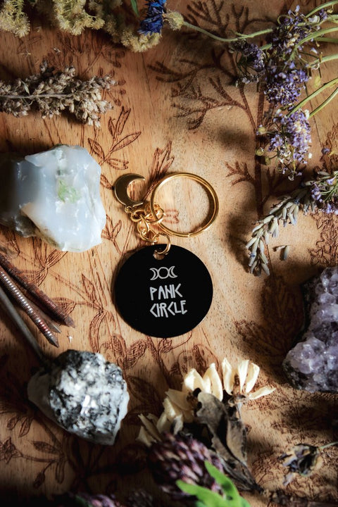 Ein Schlüsselanhänger in Schwarz und Gold mit goldenem Mond-Charm. Auf der schwarzen Scheibe steht in weiß: Panic Circle