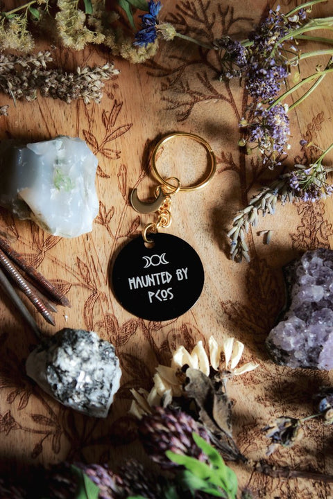 Ein Schlüsselanhänger in Schwarz und Gold mit goldenem Mond-Charm. Auf der schwarzen Scheibe steht in weiß: Haunted by PCOS